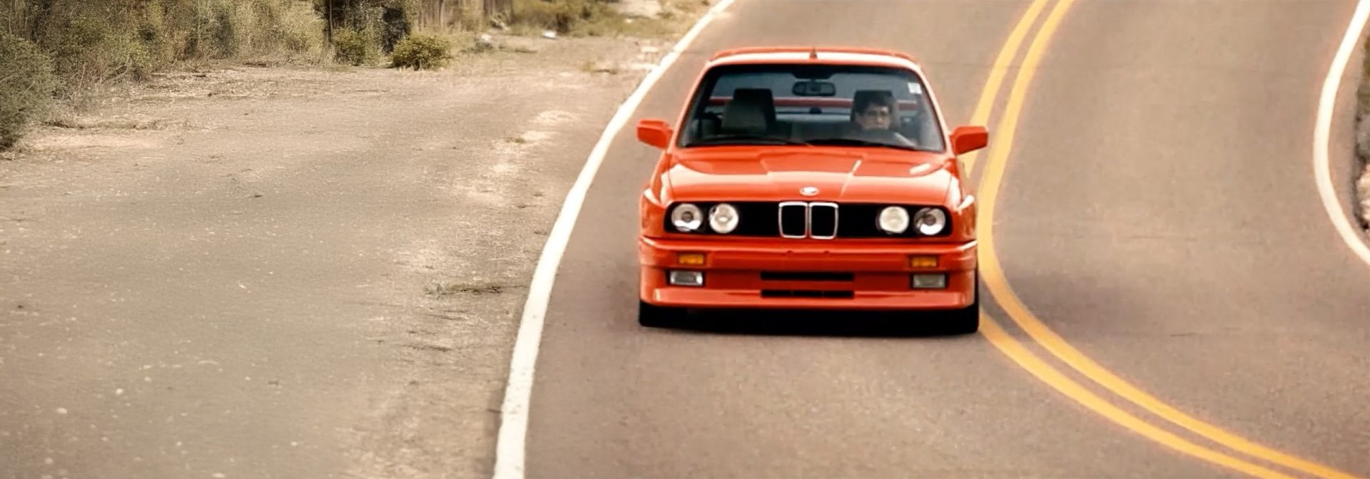 A red second-generation BMW, built between 1982 and 1994, approaches us on a country road with a double solid yellow centre line.