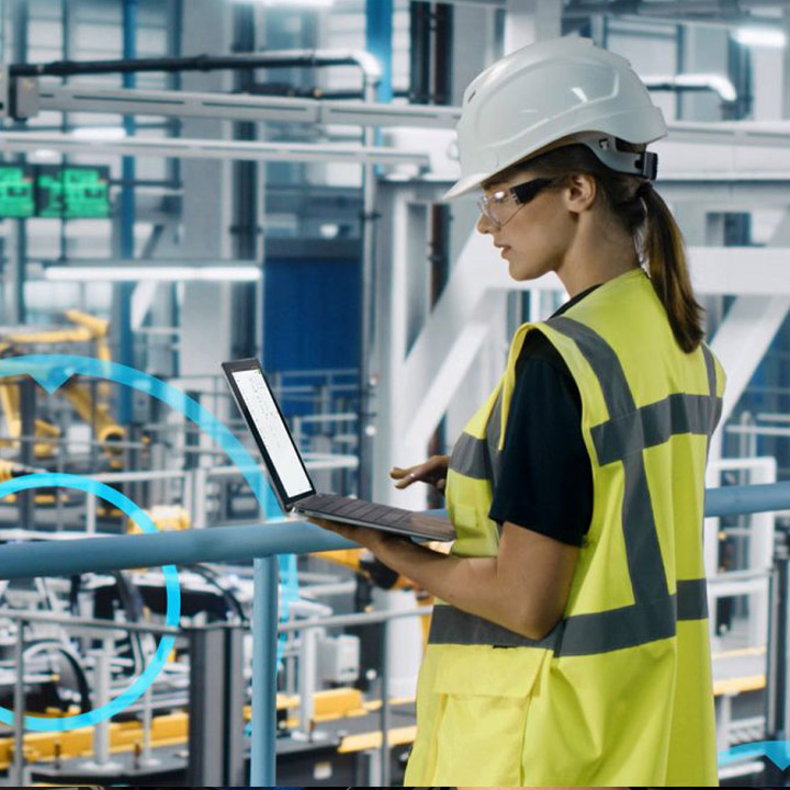 BMW employee with long brown hair, safety goggles, safety helmet and high-visibility vest enters data hands-free on a laptop.