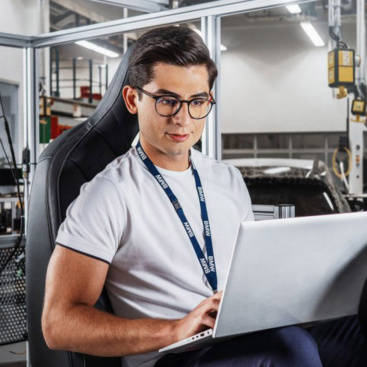 A BMW employee sits on an ergonomic car seat with his laptop open on his knees in the production area and enters data.