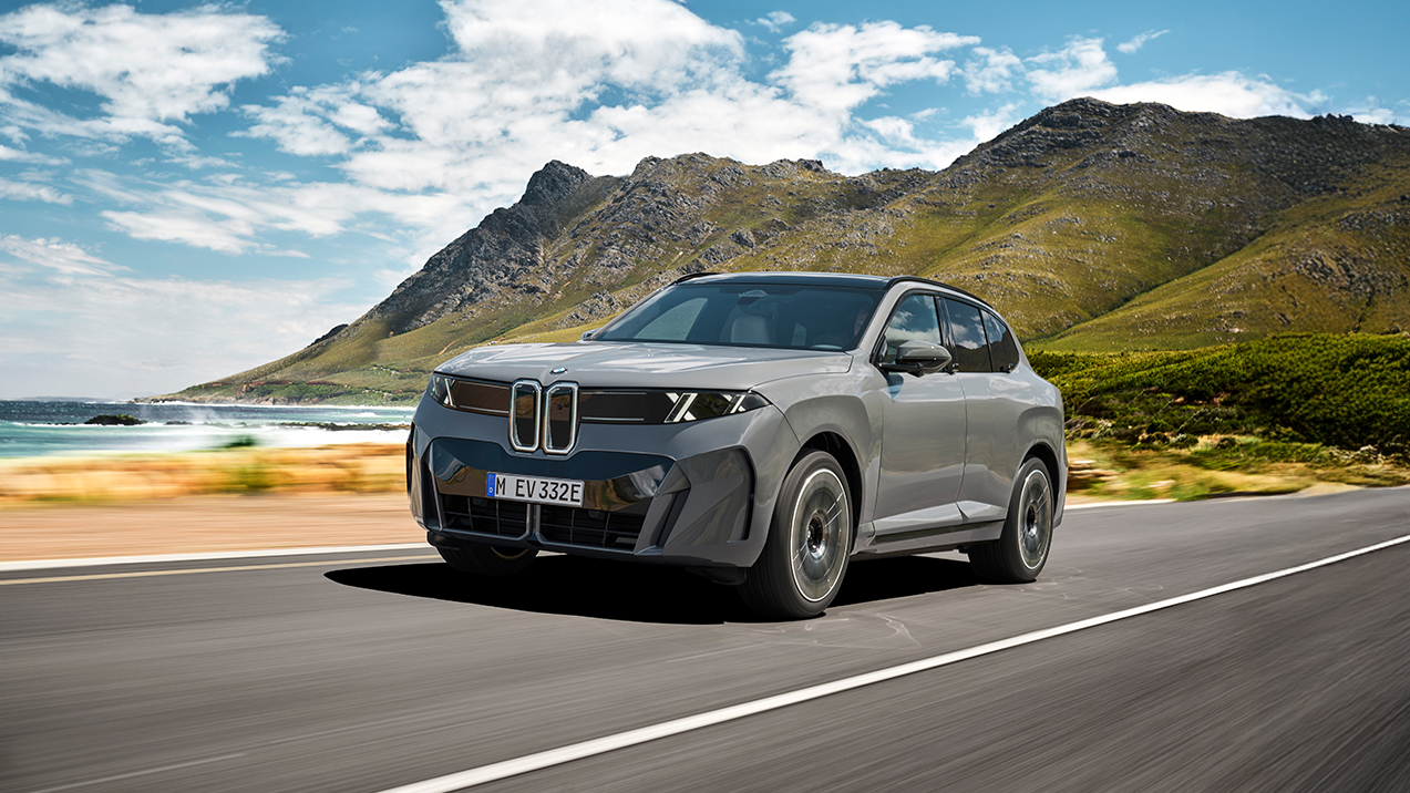 We see the new BMW ix5 driving over a street near the sea and mountains.
