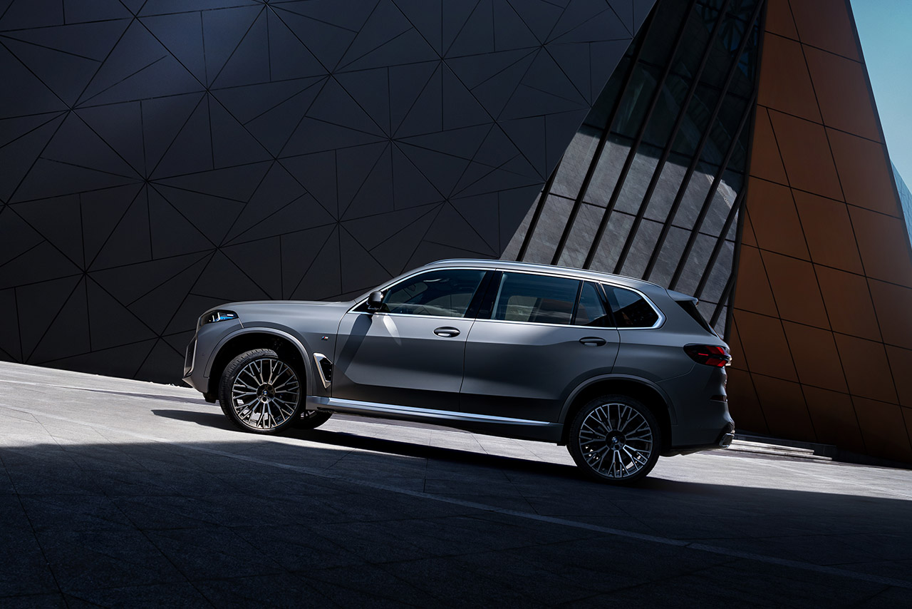 In front of a modern futuristic building, a large silver BMW SUV drives up an upward-sloping ramp.