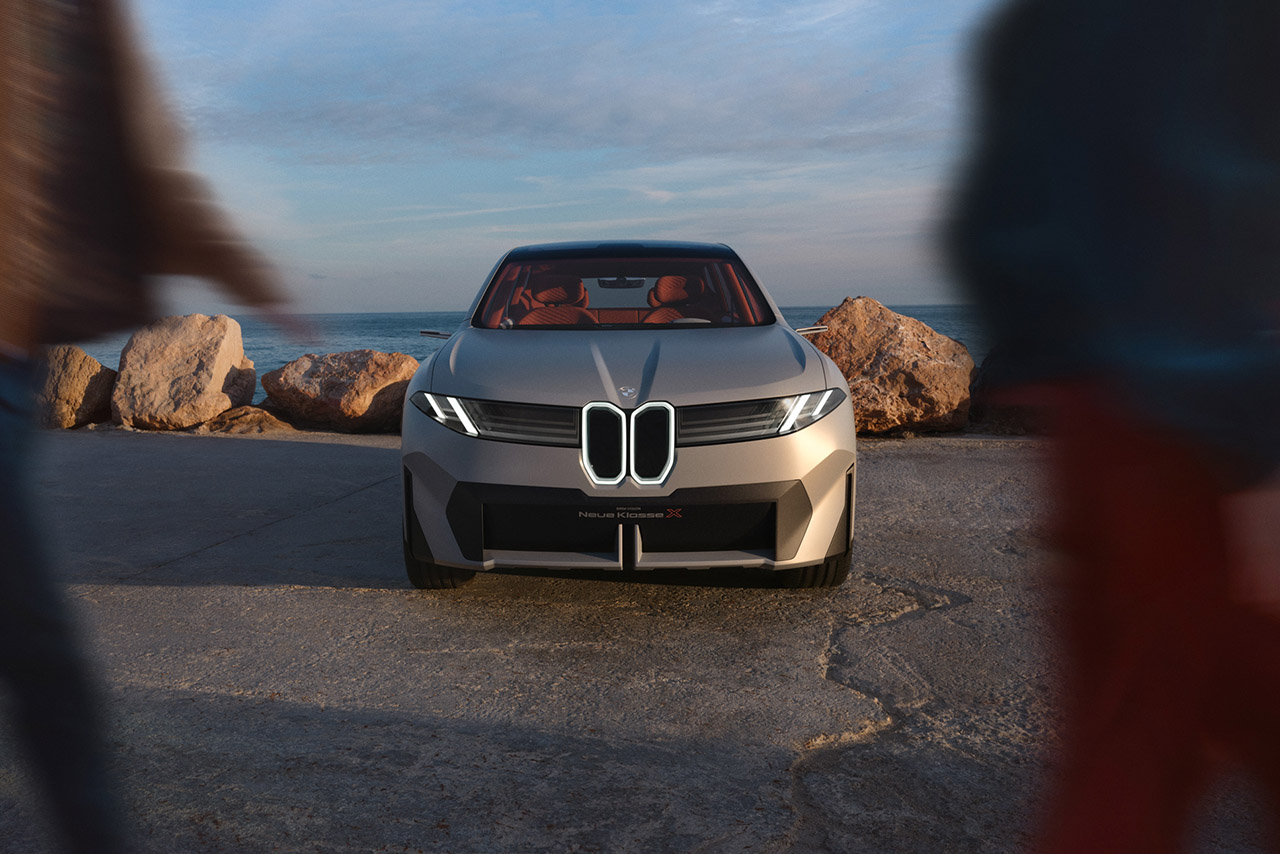 Two people looking at the BMW Neue Klasse which is parked next to the sea.