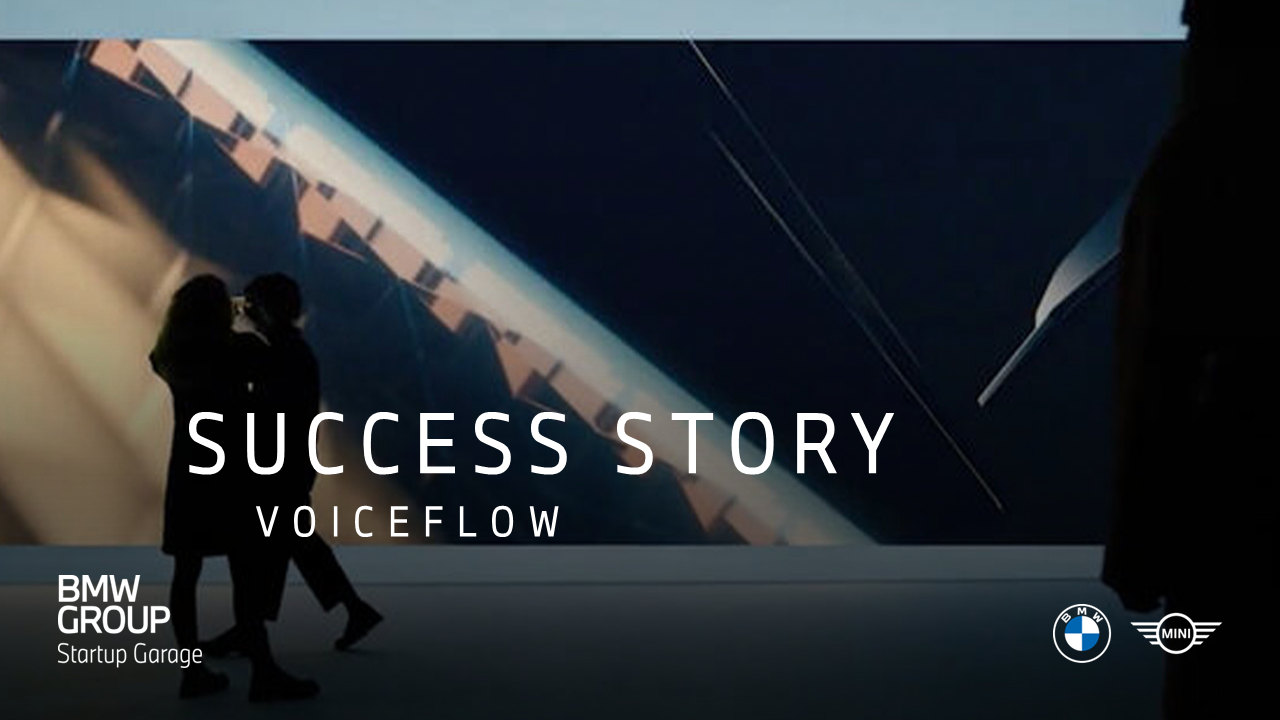 Two people stand in front of a large illuminated abstract display in a dark exhibition-like space.