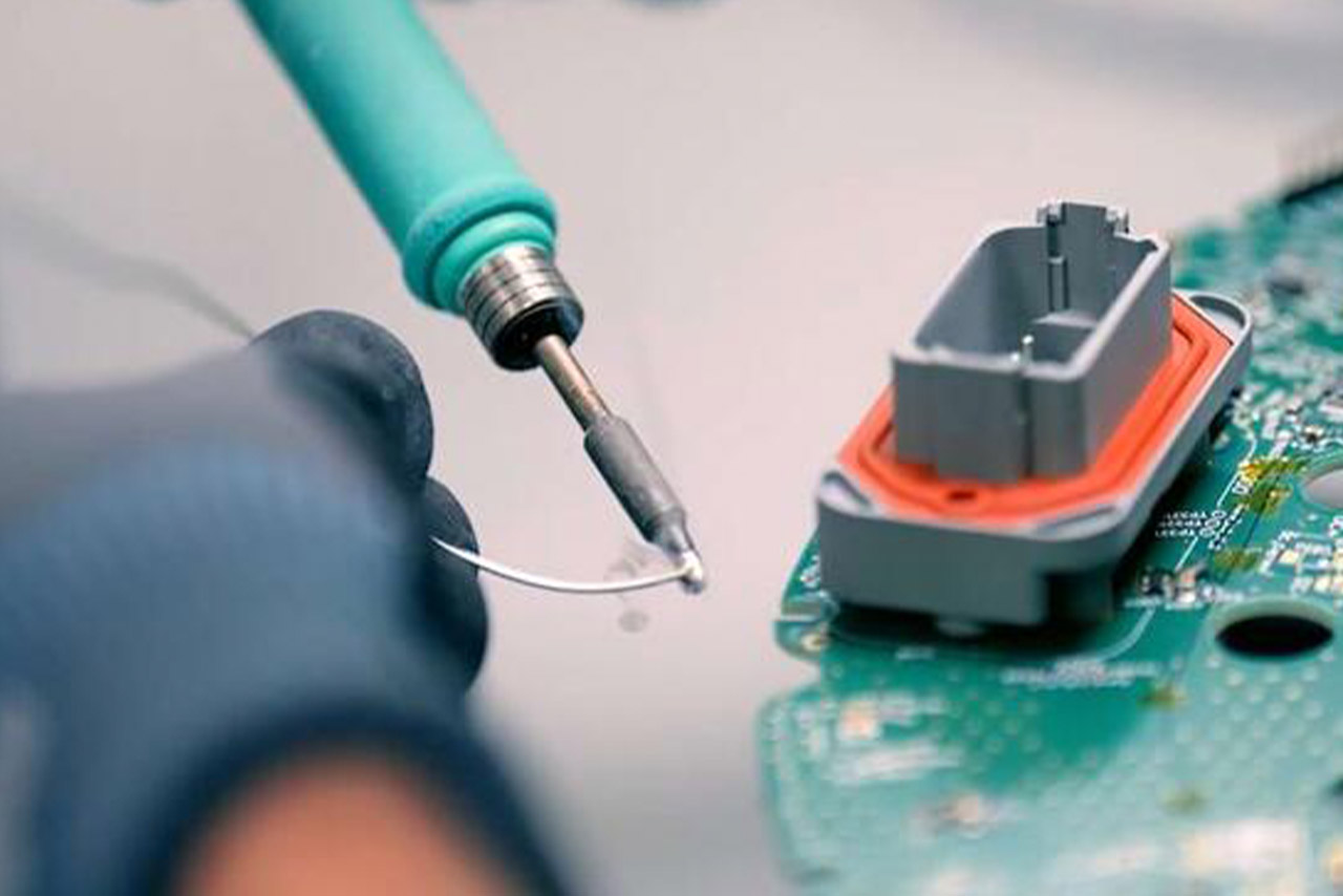 We see a green soldering iron and a hand holding a soldering wire to the tip of the device in order to attach a plug to a circuit board in the next step.